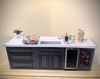 May include: A miniature kitchen island with a white countertop and dark gray cabinets. It features a black dish rack with a copper pot, a silver coffee pot, a white pitcher, and a box of Kellogg's Corn Flakes. A small potted plant sits on the left.