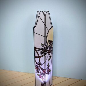 May include: A tall, white stained glass lamp with a floral design. The lamp has a white background with black outlines and pink and white flowers. The lamp is lit from the inside, casting a warm glow.