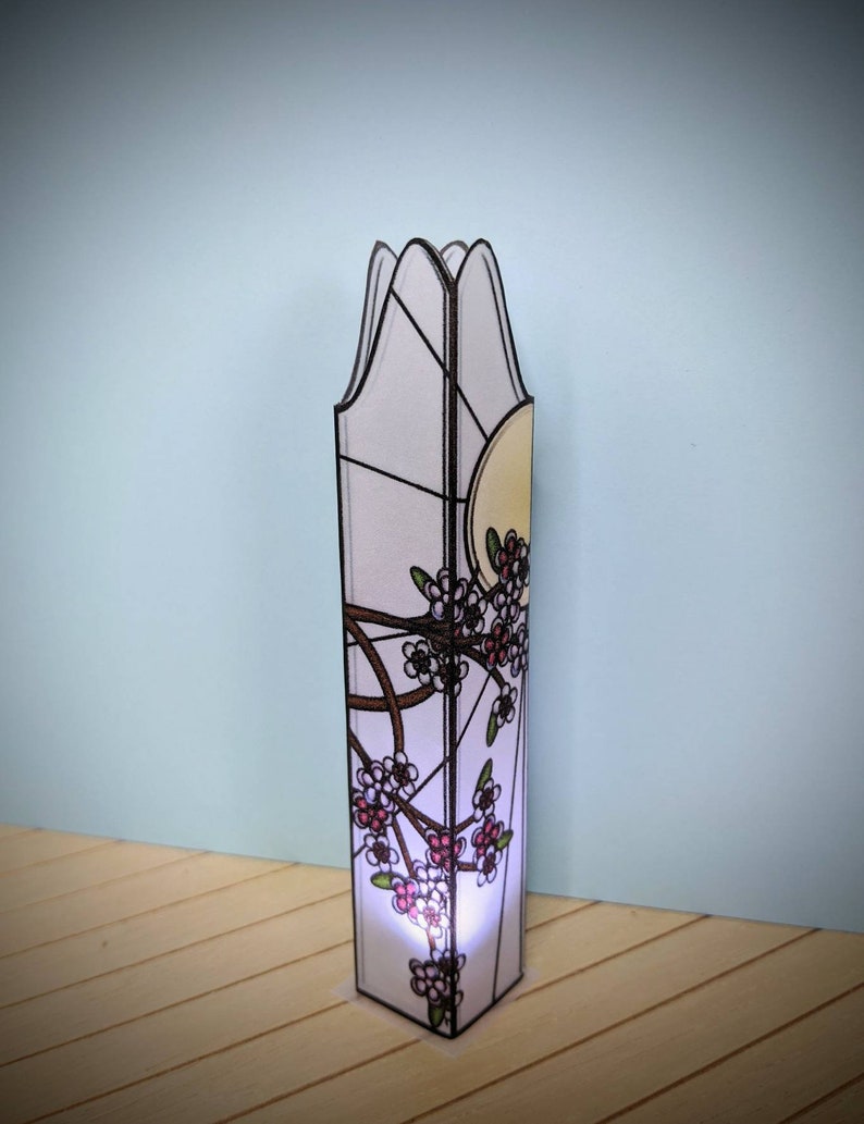 May include: A tall, white stained glass lamp with a floral design. The lamp has a white background with black outlines and pink and white flowers. The lamp is lit from the inside, casting a warm glow.