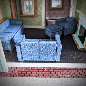 May include: A dollhouse living room with a blue patterned sofa, two armchairs, and an ottoman. The room has a brick fireplace and two windows.