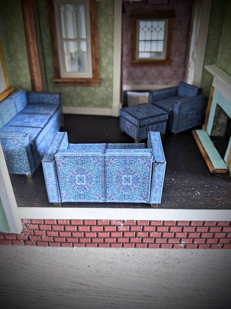 May include: A dollhouse living room with a blue patterned sofa, two armchairs, and an ottoman. The room has a brick fireplace and two windows.
