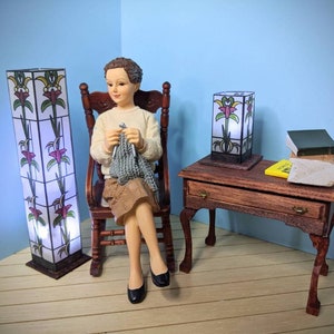May include: A doll sitting in a rocking chair knitting with two stained glass lamps and a small wooden table with books on top.