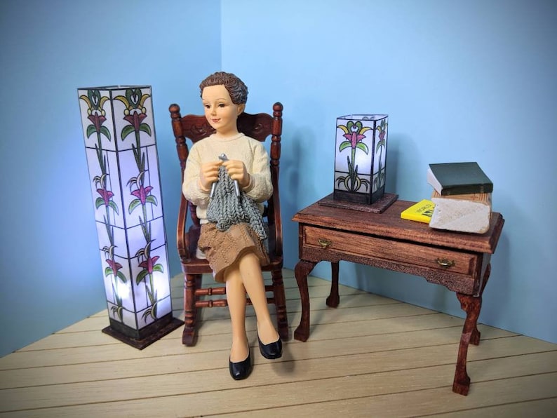 May include: A doll sitting in a rocking chair knitting with two stained glass lamps and a small wooden table with books on top.