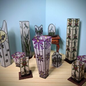 May include: A collection of eight stained glass lamps with floral and dragonfly designs. The lamps are in various shapes and sizes, and they are all made of clear glass with colorful accents. The lamps are arranged on a wooden surface.