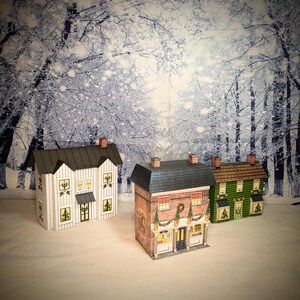 May include: Three paper model houses decorated for Christmas. The houses are white, brick, and green. The white house has a black roof and white trim. The brick house has a black roof and a red door. The green house has a brown roof and a green door. The houses are set against a snowy forest backdrop.
