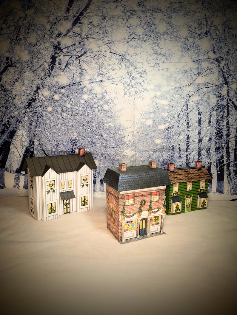 May include: Three paper model houses decorated for Christmas. The houses are white, brick, and green. The white house has a black roof and white trim. The brick house has a black roof and a red door. The green house has a brown roof and a green door. The houses are set against a snowy forest backdrop.