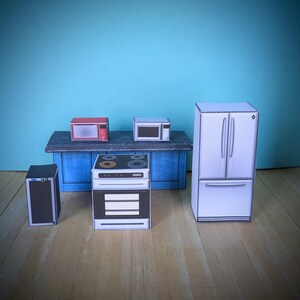 Pode incluir: Um conjunto de eletrodom&eacute;sticos de cozinha em miniatura, incluindo um forno de micro-ondas vermelho, um forno de micro-ondas branco, um forno branco com um fog&atilde;o preto, uma geladeira branca e um alto-falante preto. Os eletrodom&eacute;sticos est&atilde;o dispostos em uma bancada azul com um piso de madeira.