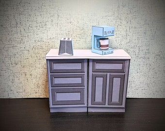 May include: A miniature kitchen set with a coffee maker and a toaster on a countertop with two cabinets. The coffee maker is light blue with a brown coffee cup. The toaster is black. The cabinets are dark brown with three drawers and two doors.