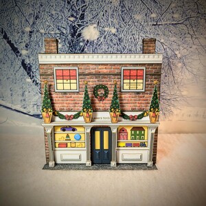 May include: A papercraft model of a toy store decorated for Christmas. The store is brick with white trim and has a sign that reads "Lamb's Toys". The windows are decorated with toys and the store is adorned with a wreath and garland.