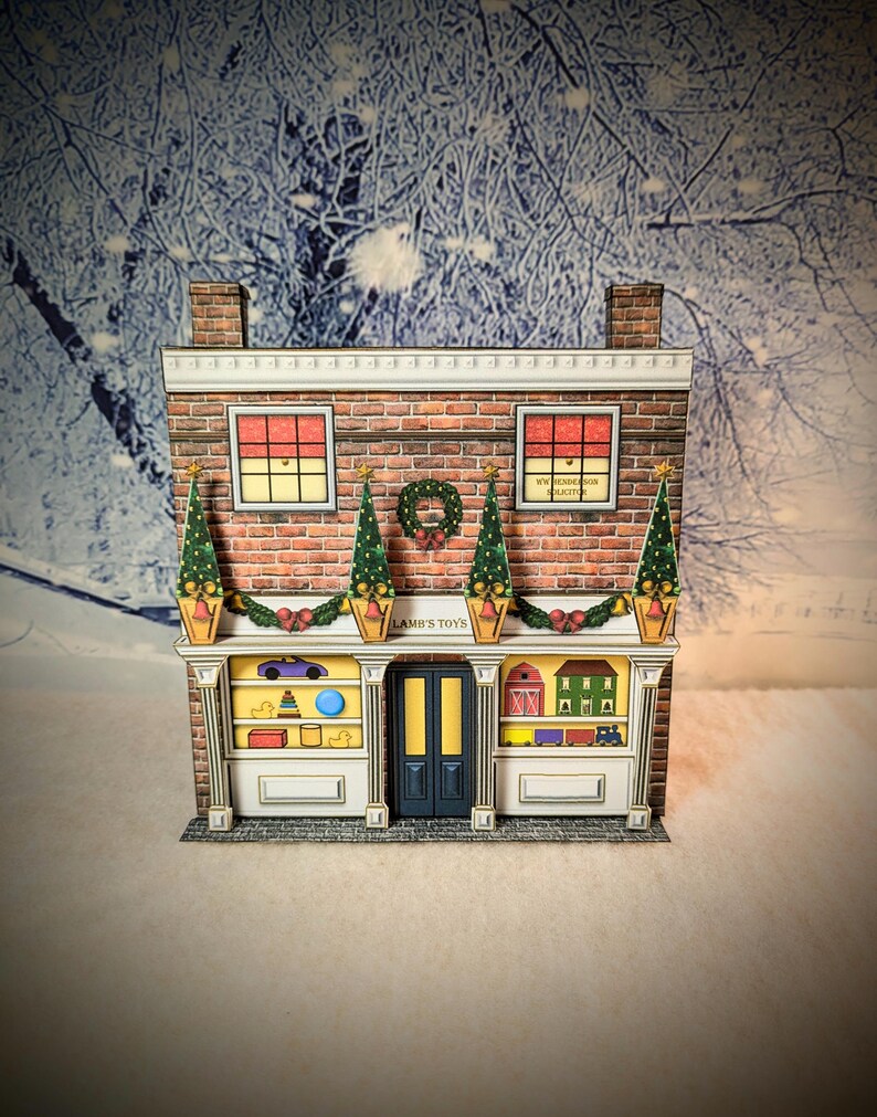 May include: A papercraft model of a toy store decorated for Christmas. The store is brick with white trim and has a sign that reads "Lamb's Toys". The windows are decorated with toys and the store is adorned with a wreath and garland.