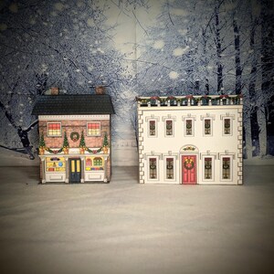 May include: Two paper model Christmas houses, one with a red door and a wreath, the other with a blue door and a sign that says "Candy Shop".