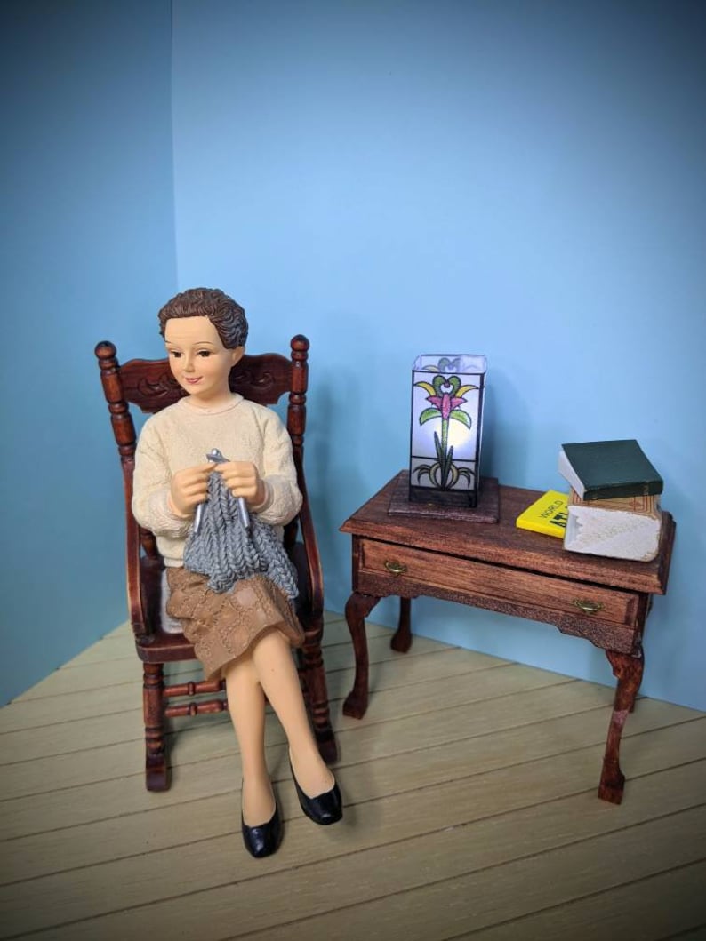 May include: A miniature doll sitting in a rocking chair, knitting with gray yarn. The doll is wearing a beige sweater and a brown skirt. A small wooden table with a stained glass lamp and books is in the background.