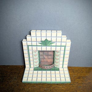 Könnte beinhalten: Ein Miniatur-Papiermodell eines Kamins mit einer gekachelten Umrandung. Die Fliesen sind cremefarben mit grünen Akzenten und einem floralen Design. Der Kamin hat einen braunen Ziegelinnenraum und einen Metallrost.