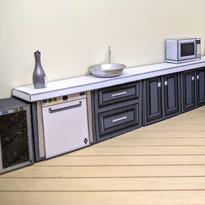 Pode incluir: Um conjunto de cozinha em miniatura com uma bancada branca, uma m&aacute;quina de lavar lou&ccedil;a branca, um refrigerador de vinho preto, uma pia de prata e arm&aacute;rios cinzentos. O conjunto inclui um forno de microondas e uma garrafa decorativa.