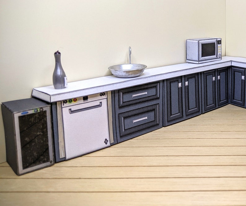 Pode incluir: Um conjunto de cozinha em miniatura com uma bancada branca, uma m&aacute;quina de lavar lou&ccedil;a branca, um refrigerador de vinho preto, uma pia de prata e arm&aacute;rios cinzentos. O conjunto inclui um forno de microondas e uma garrafa decorativa.