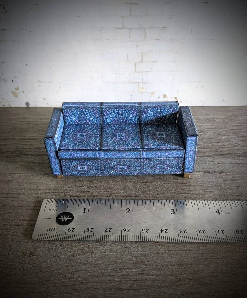 May include: A blue patterned paper model of a sofa, approximately 3 inches long, sitting on a wooden surface with a ruler for scale.