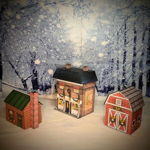 May include: Three papercraft Christmas buildings, a red barn with a wreath on the door, a red brick house with a green roof and a sign that says "Christmas Shop", and a log cabin with a green roof.
