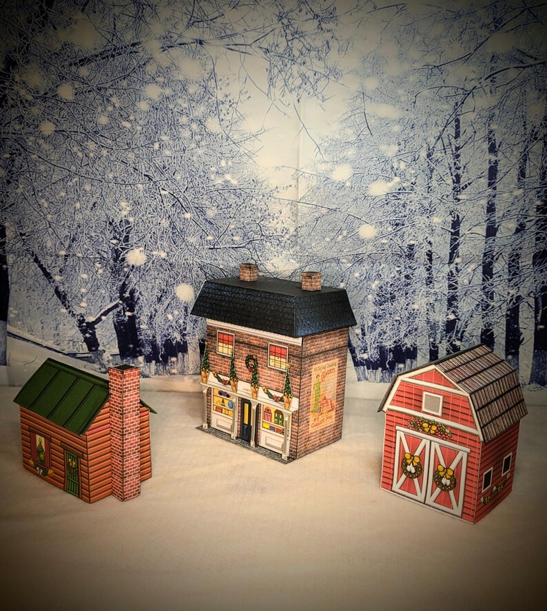 May include: Three papercraft Christmas buildings, a red barn with a wreath on the door, a red brick house with a green roof and a sign that says "Christmas Shop", and a log cabin with a green roof.