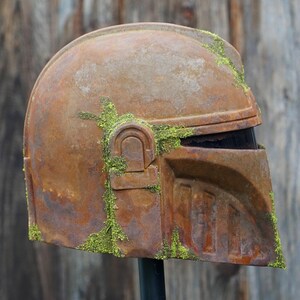 May include: A weathered, rusty brown helmet with a visor, covered in patches of green moss. The helmet has a distinct, angular design, suggesting a sci-fi or cosplay theme. The helmet is mounted on a black stand.