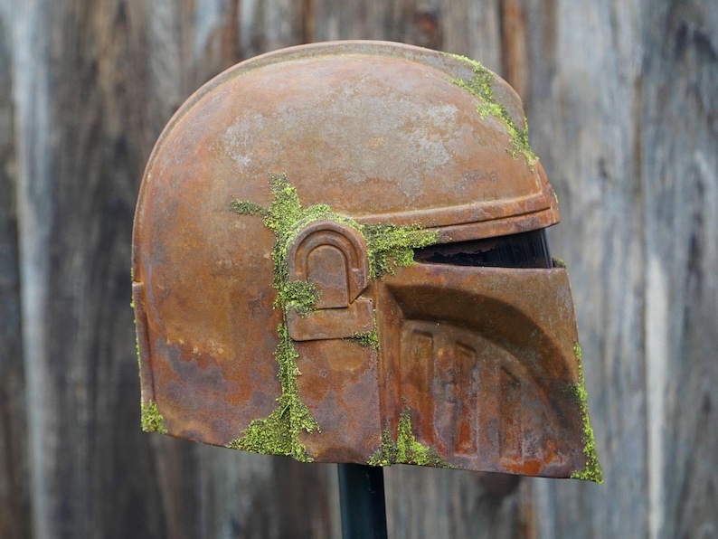 May include: A weathered, rusty brown helmet with a visor, covered in patches of green moss. The helmet has a distinct, angular design, suggesting a sci-fi or cosplay theme. The helmet is mounted on a black stand.