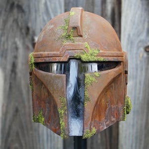 May include: A weathered, brown Mandalorian helmet with a distressed finish and green moss accents. The helmet features a T-shaped visor and is likely a cosplay or collectible item. The overall aesthetic suggests a battle-worn appearance.