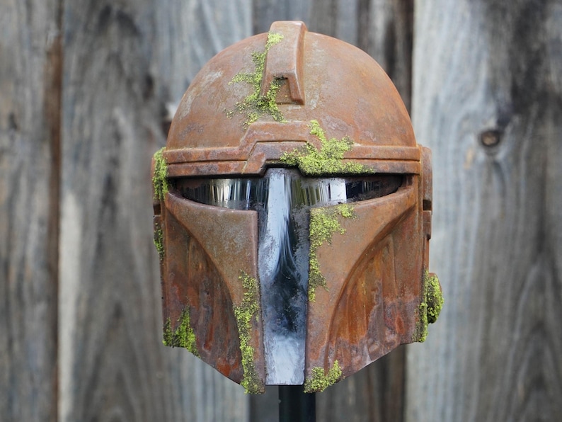 May include: A weathered, brown Mandalorian helmet with a distressed finish and green moss accents. The helmet features a T-shaped visor and is likely a cosplay or collectible item. The overall aesthetic suggests a battle-worn appearance.