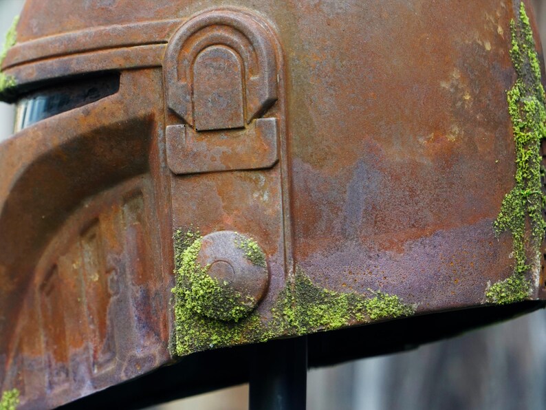 May include: Close-up of a weathered, rusty brown helmet with intricate details. Patches of green moss grow on the surface, adding texture. The helmet has a visor and a circular button detail. The overall aesthetic suggests a vintage or post-apocalyptic theme.