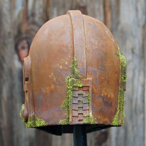 May include: A weathered, rusty metal helmet with a domed top and a handle. The helmet has rectangular side features and is partially covered in green moss. The background is a blurred wooden surface.