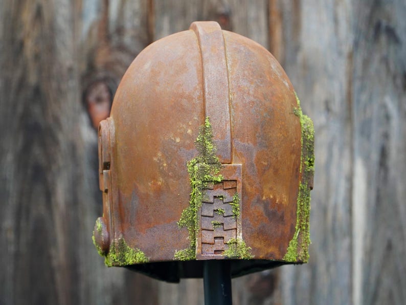 May include: A weathered, rusty metal helmet with a domed top and a handle. The helmet has rectangular side features and is partially covered in green moss. The background is a blurred wooden surface.