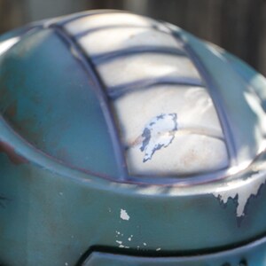 K&ouml;nnte beinhalten: Nahaufnahme eines verwitterten, teal-farbenen Helms. Der Helm hat ein segmentiertes Design mit dunklen Linien und sichtbaren Abnutzungserscheinungen, einschlie&szlig;lich abbl&auml;tternder Farbe und rost&auml;hnlicher Verf&auml;rbungen. Die Oberfl&auml;che reflektiert Sonnenlicht.