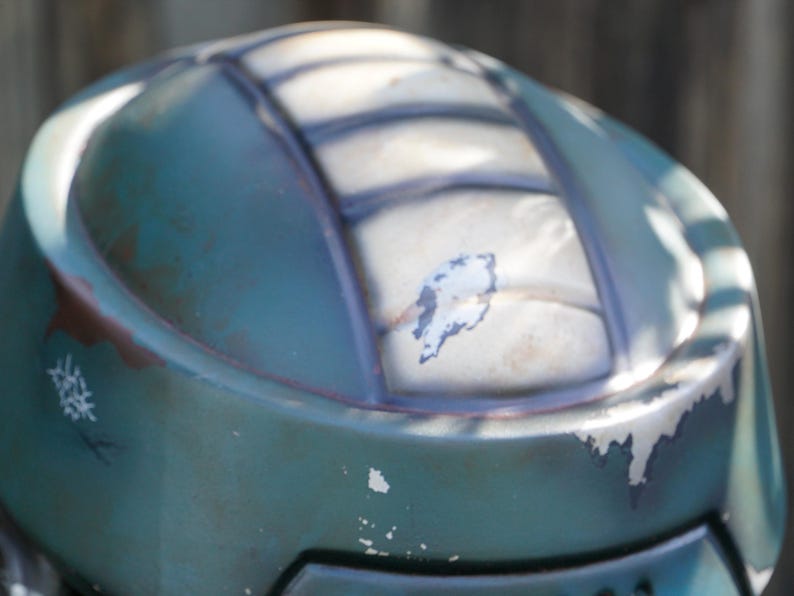 K&ouml;nnte beinhalten: Nahaufnahme eines verwitterten, teal-farbenen Helms. Der Helm hat ein segmentiertes Design mit dunklen Linien und sichtbaren Abnutzungserscheinungen, einschlie&szlig;lich abbl&auml;tternder Farbe und rost&auml;hnlicher Verf&auml;rbungen. Die Oberfl&auml;che reflektiert Sonnenlicht.