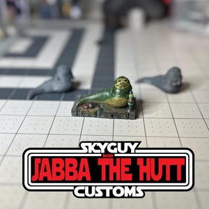 May include: A green and brown painted miniature figurine of Jabba the Hutt from Star Wars, sitting on a small base. The figurine is on a white gridded surface. The text "Skyguy Mini Jabba the Hutt Customs" is visible in the lower part of the image.