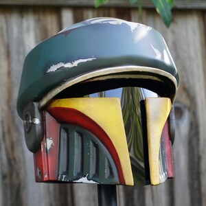 K&ouml;nnte beinhalten: Verwitterter Helm in Gr&uuml;n, Gelb und Rot. Der Helm hat ein Visier und weist Gebrauchsspuren auf, mit einem verblassten Farbeffekt. Das Design erinnert an eine beliebte Science-Fiction-Figur, wahrscheinlich f&uuml;r Cosplay oder Ausstellungen.