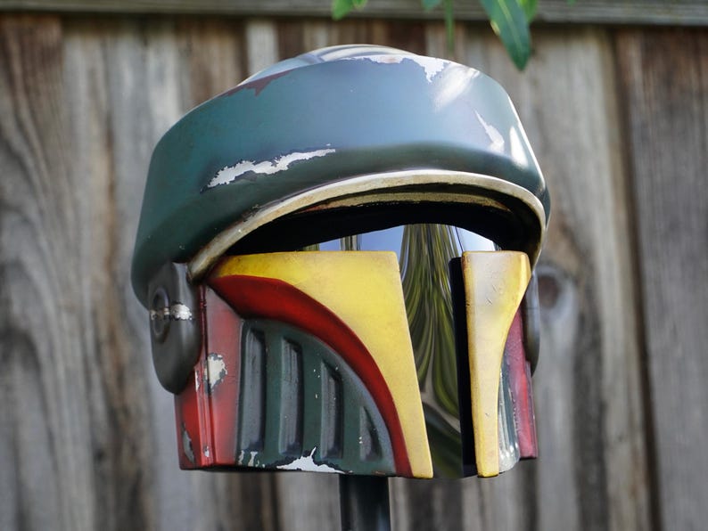 K&ouml;nnte beinhalten: Verwitterter Helm in Gr&uuml;n, Gelb und Rot. Der Helm hat ein Visier und weist Gebrauchsspuren auf, mit einem verblassten Farbeffekt. Das Design erinnert an eine beliebte Science-Fiction-Figur, wahrscheinlich f&uuml;r Cosplay oder Ausstellungen.