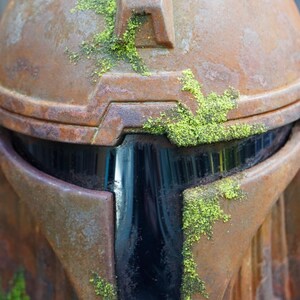 May include: Close-up of a weathered, rusty brown helmet with a dark visor. Patches of bright green moss grow on the helmet's surface, adding a natural contrast to the metallic texture. The helmet's design suggests a sci-fi or fantasy theme.