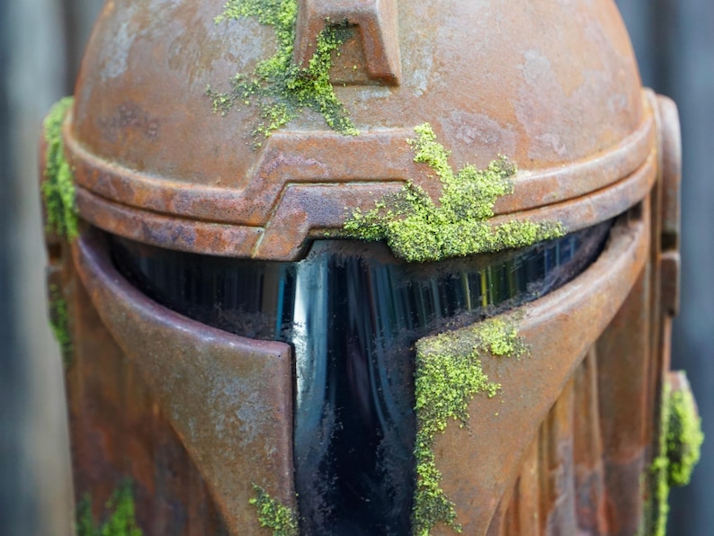 May include: Close-up of a weathered, rusty brown helmet with a dark visor. Patches of bright green moss grow on the helmet's surface, adding a natural contrast to the metallic texture. The helmet's design suggests a sci-fi or fantasy theme.