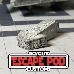 Puede incluir: Un modelo en miniatura de una cápsula de escape gris metálico con paneles detallados y características mecánicas. El modelo está etiquetado "SKYGUY MINI ESCAPE POD CUSTOMS" en un gráfico rojo y blanco. La cápsula está sobre un fondo de cuadrícula gris y blanco.