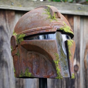 May include: A weathered, brown helmet with a visor, covered in patches of green moss. The helmet has a rugged, metallic appearance, suggesting a sci-fi or fantasy theme. The design features angular shapes and a prominent brow ridge.