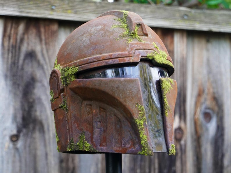 May include: A weathered, brown helmet with a visor, covered in patches of green moss. The helmet has a rugged, metallic appearance, suggesting a sci-fi or fantasy theme. The design features angular shapes and a prominent brow ridge.