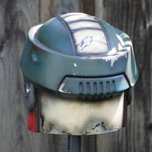 K&ouml;nnte beinhalten: Ein verwitterter, teal-farbener Helm mit wei&szlig;en und grauen Details. Der Helm hat eine abgerundete Oberseite mit rechteckigen L&uuml;ftungsschlitzen und einem roten Akzent. Die Lackierung weist Gebrauchsspuren auf und wirkt dadurch vintage.