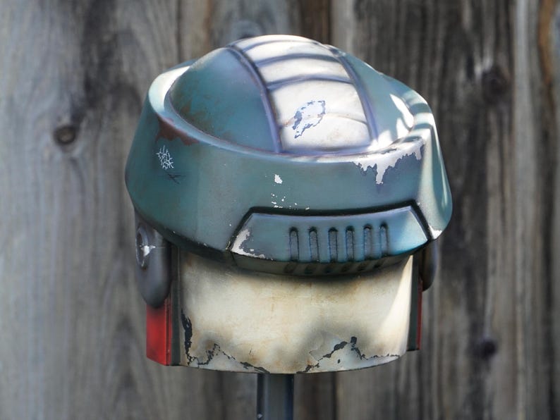 K&ouml;nnte beinhalten: Ein verwitterter, teal-farbener Helm mit wei&szlig;en und grauen Details. Der Helm hat eine abgerundete Oberseite mit rechteckigen L&uuml;ftungsschlitzen und einem roten Akzent. Die Lackierung weist Gebrauchsspuren auf und wirkt dadurch vintage.