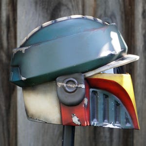 K&ouml;nnte beinhalten: Ein verwitterter Helm in Gr&uuml;n-, Gelb-, Rot- und Off-White-T&ouml;nen. Der Helm hat ein Visier und eine abgerundete Oberseite mit sichtbaren Gebrauchsspuren, was auf eine kampferprobte &Auml;sthetik hindeutet. Das Design erinnert an eine Weltraumfigur.