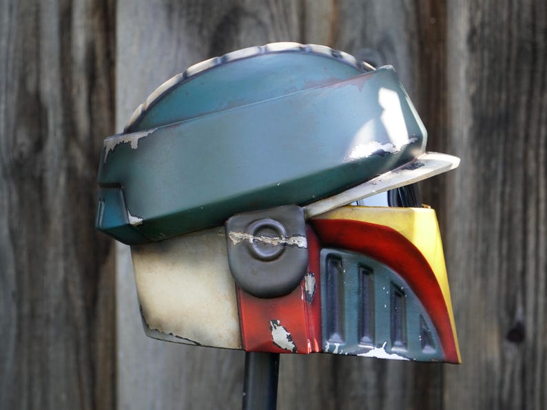 K&ouml;nnte beinhalten: Ein verwitterter Helm in Gr&uuml;n-, Gelb-, Rot- und Off-White-T&ouml;nen. Der Helm hat ein Visier und eine abgerundete Oberseite mit sichtbaren Gebrauchsspuren, was auf eine kampferprobte &Auml;sthetik hindeutet. Das Design erinnert an eine Weltraumfigur.
