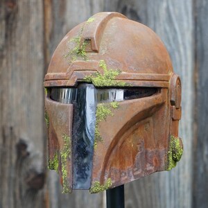 May include: A weathered, brown Mandalorian helmet with a distressed, rusty appearance. The helmet features a visor and is adorned with patches of green moss, giving it a battle-worn look. The helmet is displayed against a wooden background.