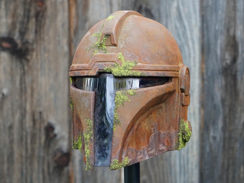 May include: A weathered, brown Mandalorian helmet with a distressed, rusty appearance. The helmet features a visor and is adorned with patches of green moss, giving it a battle-worn look. The helmet is displayed against a wooden background.