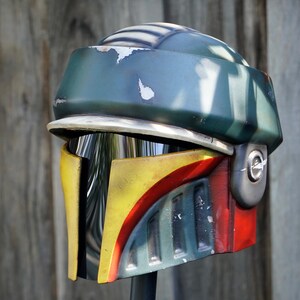 K&ouml;nnte beinhalten: Ein detaillierter, verwitterter Helm mit einem unverwechselbaren Design. Der Helm hat eine gr&uuml;ne Oberseite, eine gelbe und rote Frontplatte und ein silbernes Visier. Das Design deutet auf ein Science-Fiction- oder Cosplay-Thema hin.