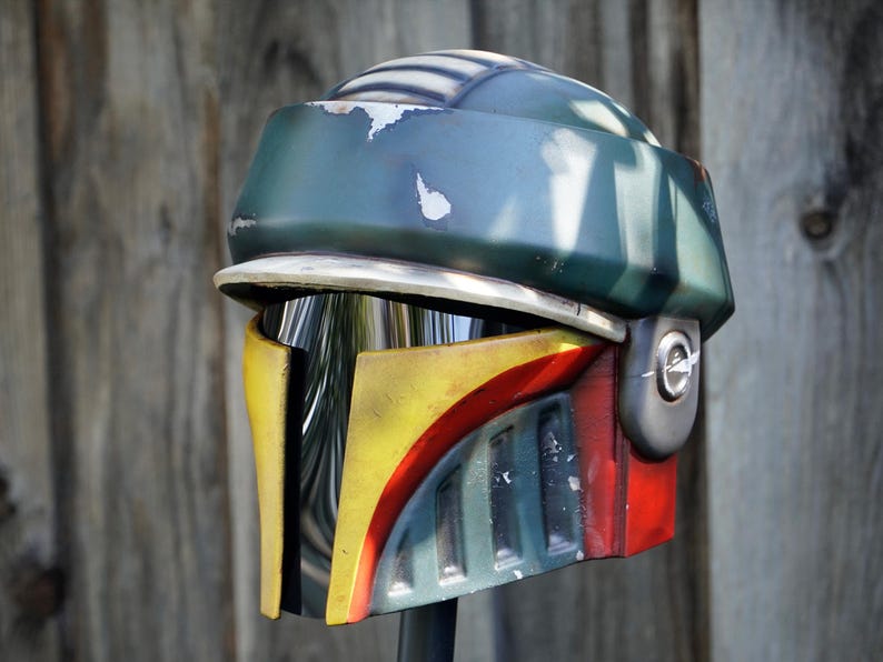 K&ouml;nnte beinhalten: Ein detaillierter, verwitterter Helm mit einem unverwechselbaren Design. Der Helm hat eine gr&uuml;ne Oberseite, eine gelbe und rote Frontplatte und ein silbernes Visier. Das Design deutet auf ein Science-Fiction- oder Cosplay-Thema hin.