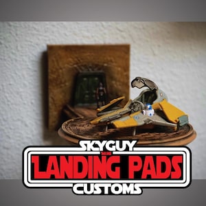 May include: A miniature yellow and gray spacecraft model with a detailed cockpit, displayed on a wooden base. The model is set against a backdrop of a miniature scene with a figure. The text "SKYGUY MINI LANDING PADS CUSTOMS" is displayed at the bottom.
