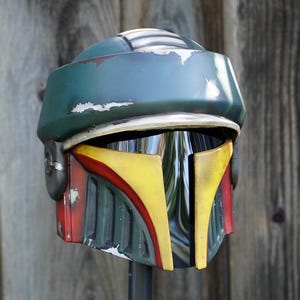 K&ouml;nnte beinhalten: Ein verwitterter Helm in Gr&uuml;n-, Gelb- und Rott&ouml;nen mit Visier. Der Helm hat eine abgerundete Oberseite und ein markantes Design mit gelben und roten Akzenten auf der Vorderseite. Es scheint sich um eine Requisite oder ein Sammlerst&uuml;ck zu handeln.