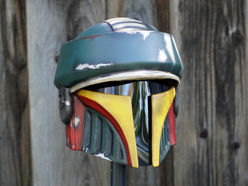 K&ouml;nnte beinhalten: Ein verwitterter Helm in Gr&uuml;n-, Gelb- und Rott&ouml;nen mit Visier. Der Helm hat eine abgerundete Oberseite und ein markantes Design mit gelben und roten Akzenten auf der Vorderseite. Es scheint sich um eine Requisite oder ein Sammlerst&uuml;ck zu handeln.